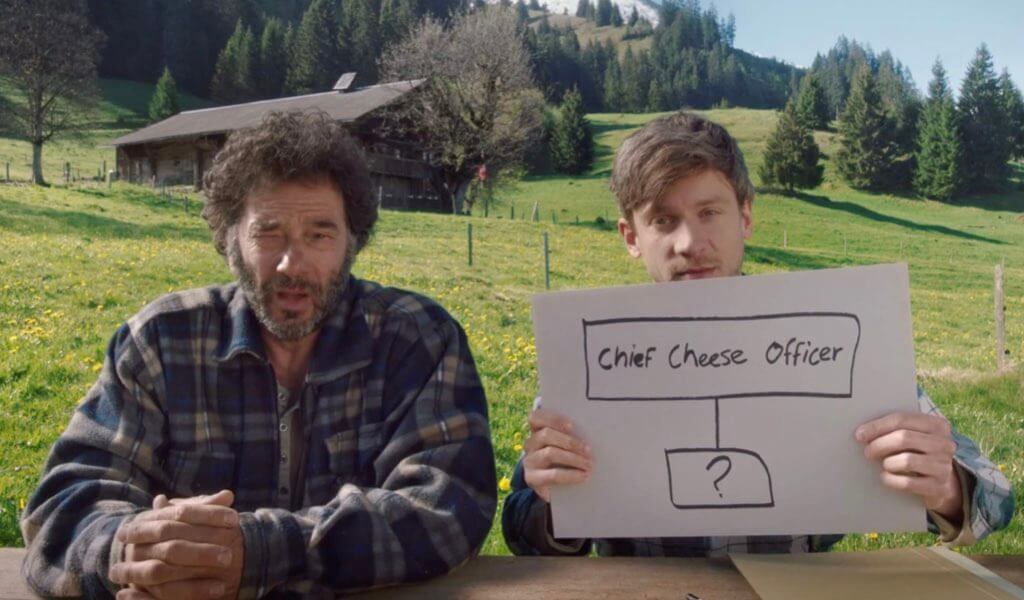 Zwei Älpler halten ein Schild. Darauf eine Art Organigramm mit dem Titel "Chief Cheese Officer"